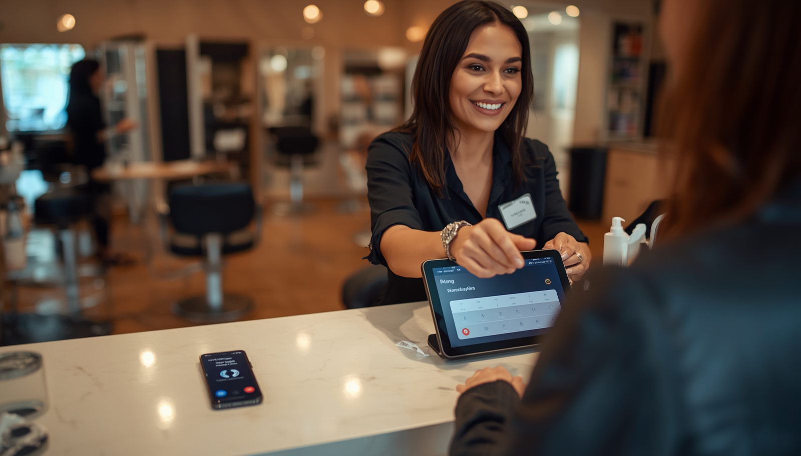 A smiling receptionist at a salon front desk, checking a client in on a tablet while a smartphone on the counter is ringing.